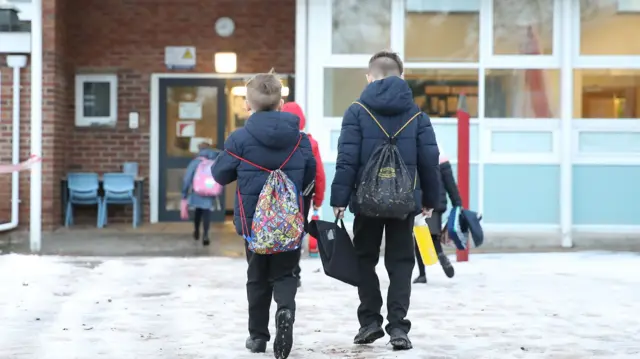 Two pupils arrive at school. They are walking to the entrance and a wearing warm coats.