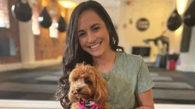 Leanne Lucas smiles at the camera as she holds a dog. An exercise studio can be seen in the background slightly blurred.