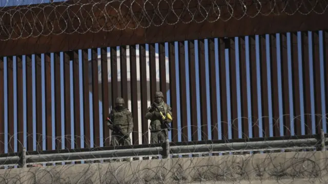 US soldiers patrol the US Mexico border - two armed soldiers stand in front of a border gate with barbed wire