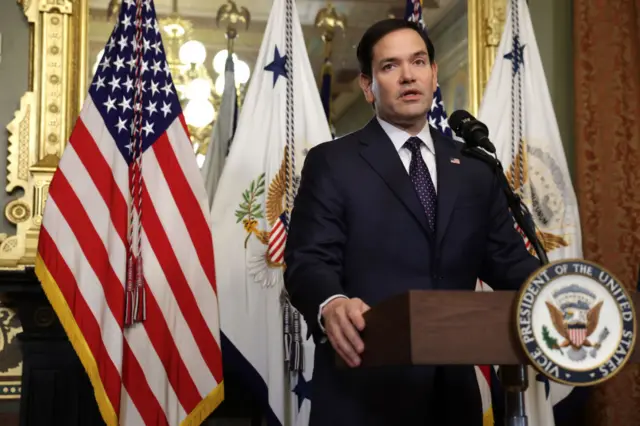 Marco Rubio stands at a lectern in front of United States of America flags, wearing a dark suit