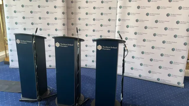 Three podiums with the logo of the Northern Ireland Executive on them, sitting in front of a background with the same logo printed on them,