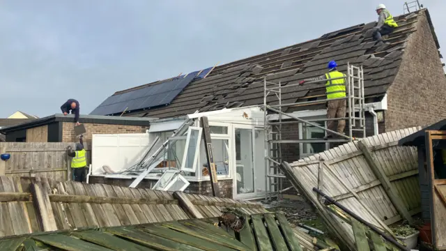 Workers are fixing damage that had occurred during a tornado. There is two people wearing a hi-vis looking to repair a roof.