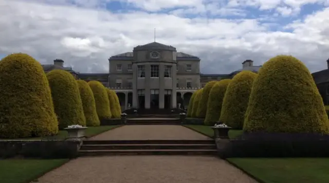 A tree-lined path leading to a grand building with pillars around the entrance