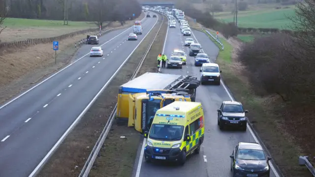 Two sides of an A road can be seen. On the left side a few vehicles are travelling on a clear road. On the right an ambulance is parked in front of an overturned lorry with a queue of cars in the right lane.