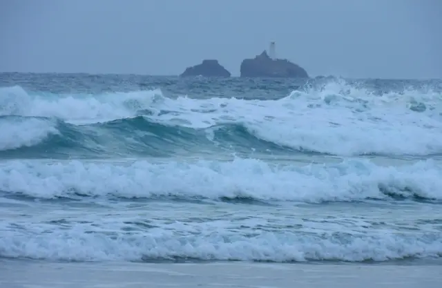 Big waves crashing on the shore at Carbis Bay. In the distance is rocks with a white lighthouse on one of them.