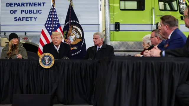 Donald Trump answers questions from the press sitting at a table covered by a black cloth, the presidential insignia at the front. Melania Trump in a black hat and green jacket sits to his right