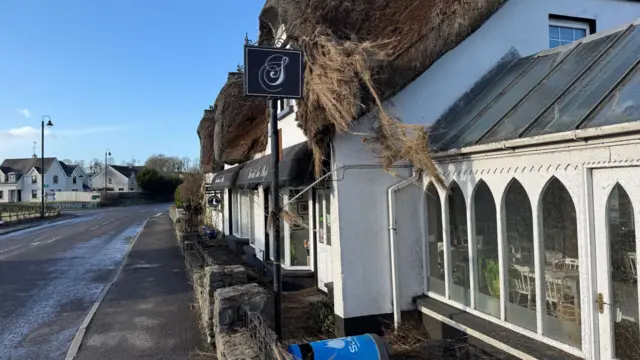 A white brick cottage with a brown thatched roof with a footpath outside, dividing it the road. The roof has been damaged and a part of it is hanging off. Their is a blue bin on it side in front of the building.