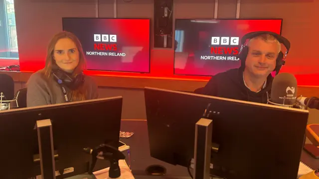 A women with ginger hair wears a grey jumper and black headphones around her neck. She sits in front of a black computer. To her left is a man with short grey hair with black headphones on his head. He wears a black jumper. Behind the two of them are two screens that read 'BBC News Northern Ireland'.