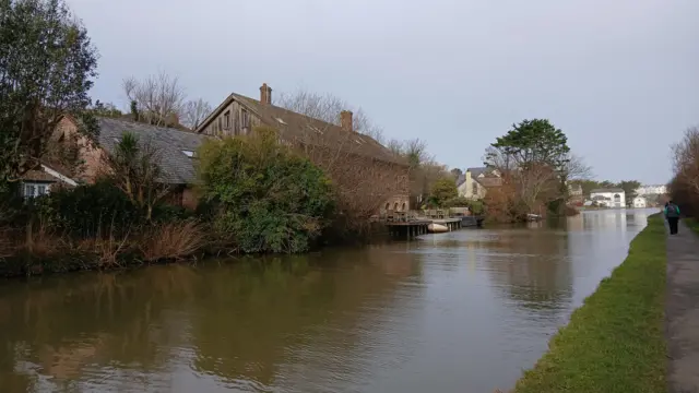 A picture of the river in Bude. The sky remains grey with people seen walking on a path next to the river