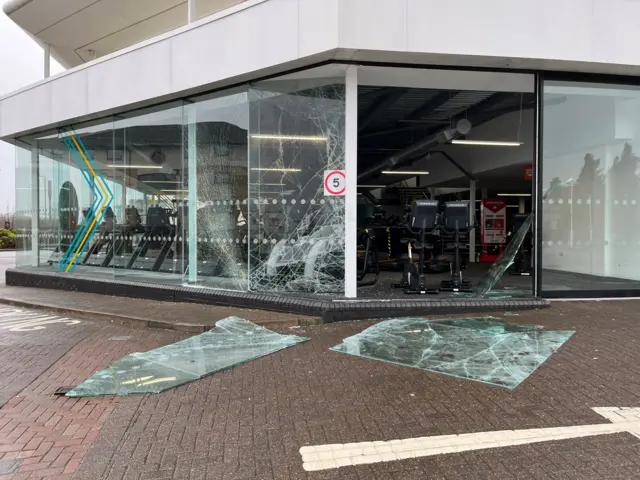 Two panes of smashed glass lie on the road. Behind them exercise bikes inside a gym are visible, as well as more shattered panes of glass which remain in the window