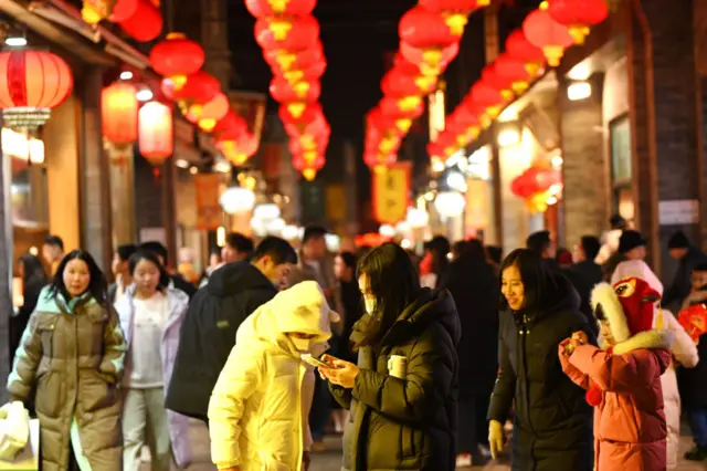 Shoppers in winter coats walking down a brightly lit street filled with red lanterns, at night