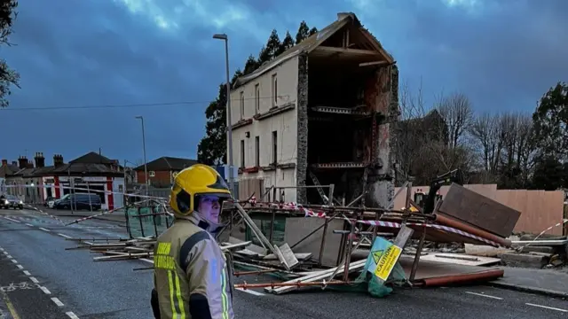 A firefighter stood on a street with a massive scaffolding structure collapsed in front of him covering the road. There's a building which is missing an entire side behind it.