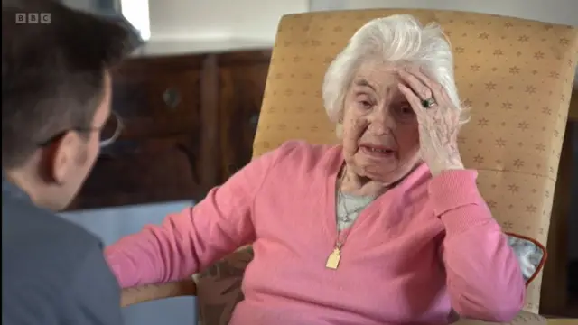 Renee Salt in a pink jumper with a grey shirt and a golden pendant around her neck sitting down on a yellow cushioned chair. Interviewer visible from the back to the left side of the frame, a wooden table blurred in the background