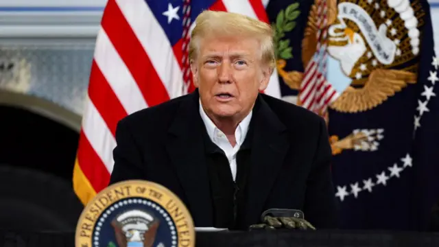 Donald Trump addresses press as he sits at table covered in black cloth. A set of leather gloves are resting on the top, a Presidential sigil at the front and a US and presidential flag in the background