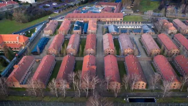 Aerial view of Auschwitz I camp with multiple red-roofed brick buildings. The main entrance's turret is recognisable in the top left of the image