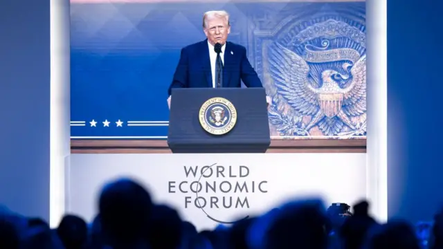 Trump onscreen at Davos, standing at a lectern with the presidential seal, speaking at the audience, whose heads can be seen at the bottom of the frame