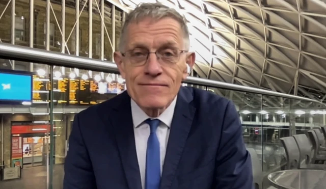 Headshot of Calder in a blue suit sitting on a balcony in a train station with the departure board behind him