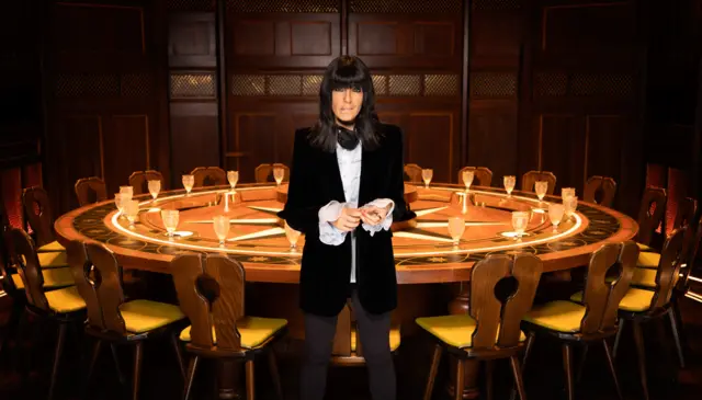 Claudia Winkleman in a black velvet blazer, sat in front of The Traitors roundtable