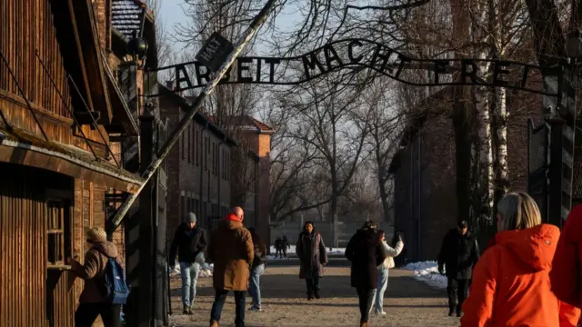 Group of tourists visit the Auschwitz I camp in POland. One woman in a brown jacket carrying a blue backpack is at the wooden ticket office speaking to someone inside. Several other people are walking across the entrance, the sign Arbeit Macht Frei (Work sets you free) at the top