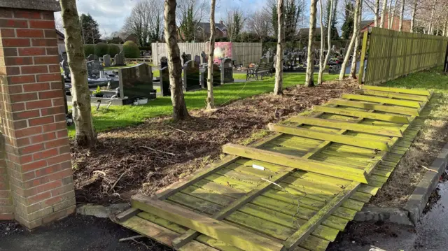 Part of a fence lies on the ground. To the left are graves in a cemetry