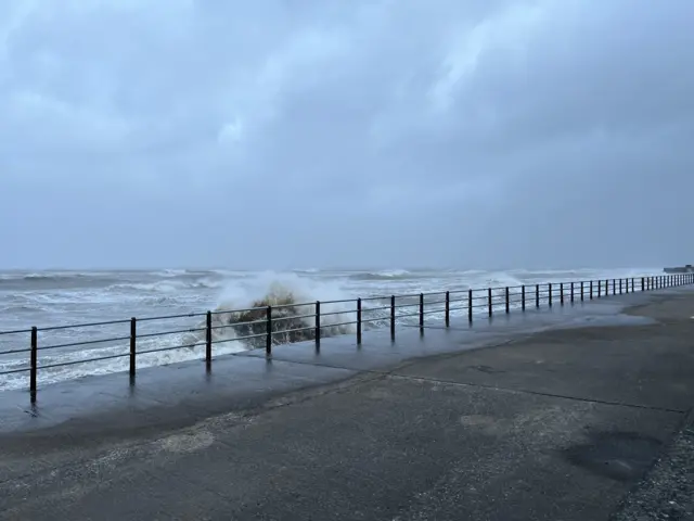 A view from a promenade next to the sea. The water is very wavy, and a large wave splashes up just before the barriers