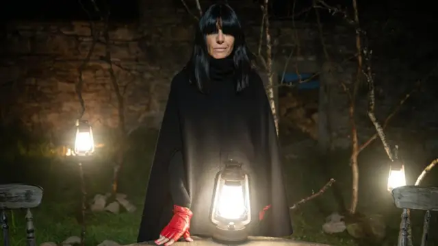 Claudia Winkleman in black cloak with a lamp on table in front of her in an outdoor setting with wall, grass and trees behind her