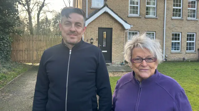 A man with short dark hair wearing a dark coloured zip up stands next to an older woman with short, blonde hair wearing rectangle frame glasses and a purple zip up jacket. They are standing in the driveway outside of the light-coloured brick house.