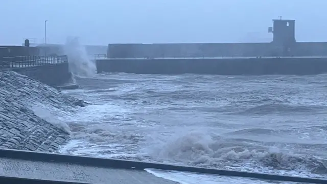 Waves crashing against the sea wall at Whitehaven Harbour. In the foreground is a jetty with rough sea in the centre of the image