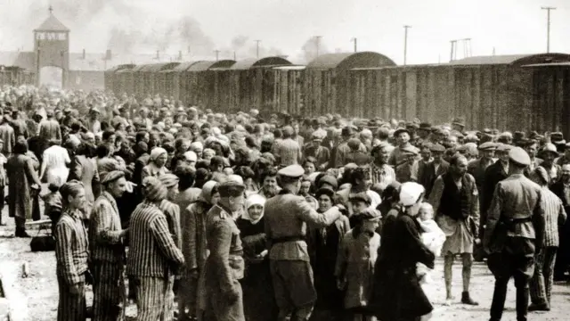 A group of Hungarian Jews in tattered clothes wait next to the rail track at Auschwitz camp, the brick turret at the end of the tracks visible in the top far left of the image. Prisoners in striped shirts and trousers can be seen waiting around as Nazi officials give orders