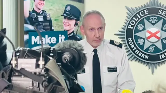 Police Davy Beck stands at a podium giving a press conference. He is wearing a police uniform and a PSNI crest is on the wall in the background.