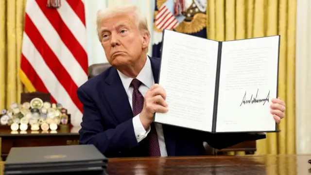 Donald Trump sits at the Oval Office desk holding a signed executive order open like a book