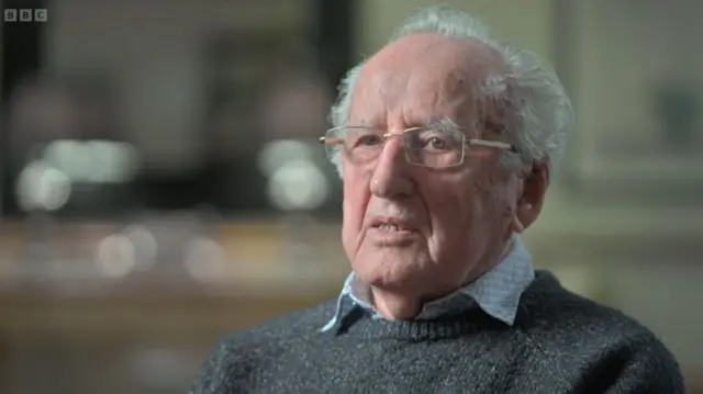 Ivor Perl in grey jumper and light blue patterned shirt underneath as he sits for the interview. The background is blurred