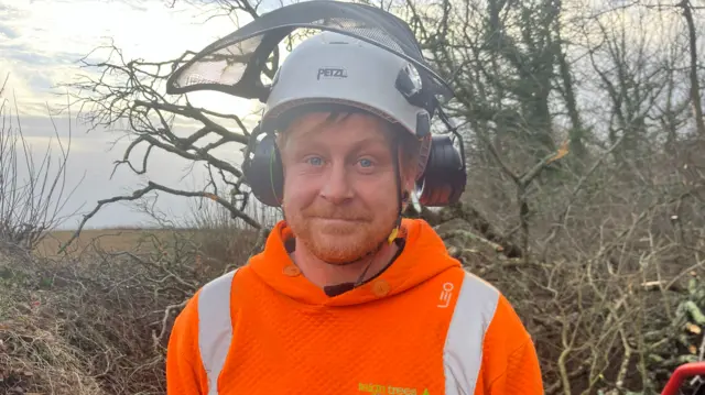 A man with ear muffs and a helmet on smiling at the camera in front of a fallen tree.