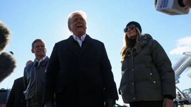 Donald Trump shot from the bottom up as he speaks to the press after arriving in Asheville. Melania Trump in a woollen hat, sunglasses and green jacket stands to his left