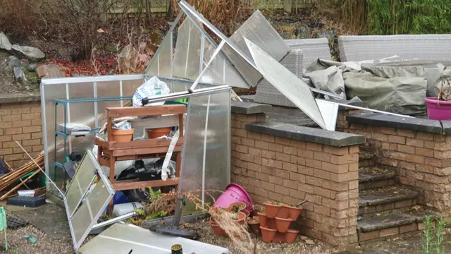 In tact panes of glass strewn - clearly once a greenhouse - are strewn across a garden. Shelves of plant pots are still visible