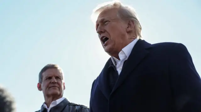 Donald Trump mid-shot as he speaks to the press. He's standing next to one North Carolina official wearing a winter coat in black and a white shirt