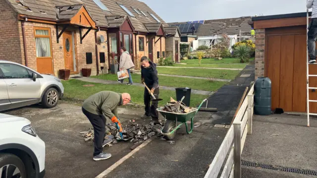 Two men are pictured using spades to move broken tiles into a wheelie barrow in front of a house.