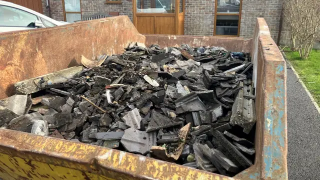 A skip is full of broken tiles. It is full of black and brown tiles in front of a brick house.