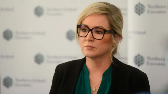 Michell O'Neill, she has shoulder length blonde hair and black-rimmed glasses and is wearing a black jacket over a green blouse