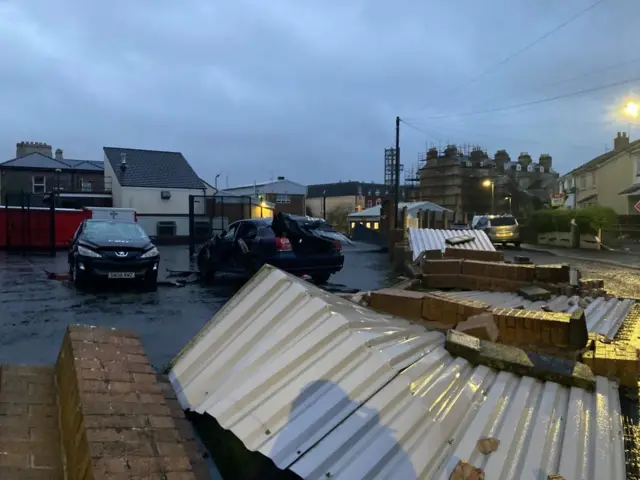 A wall and fences blown over, and cars that have been damaged and the boot is open