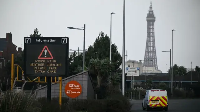 A sign on a road has a warning that says Amber Winder Weather Warning Please Take Extra Care. In the background, there is Blackpool Tower and a grey, cloudy sky.