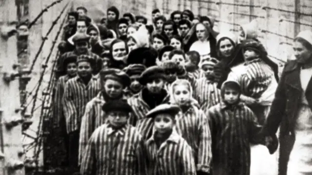 Black and white image of children survivors at Auschwitz camp after liberation. The children, wearing striped striped shirts and hats, are behind a barbed wire fence with a woman looking like a nurse to the far right of the frame holding a child's hand