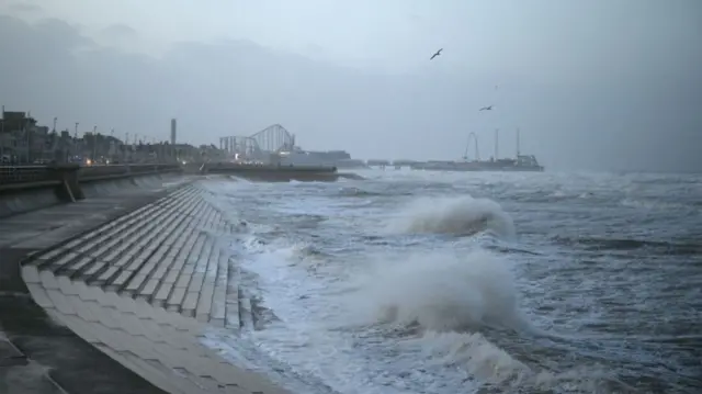 Waves at Blackpool crash onto the promenade wall. Rollercoasters at Blackpool Pleasure Beach are visible in the distance.