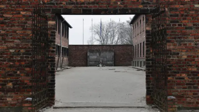 Long shot view of the death wall inside Auschwitz, in a yard on the side of Block 11. A section of the stone wall with floral tributes is in the background surrounded by red brick buildings