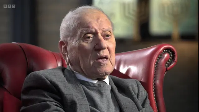 Arek Hersh in a grey jacket, grey vest and white shirt sitting down on a red leather armchair. Two golden menorahs on a windowsill are visible in the blurred background to the left of the chair