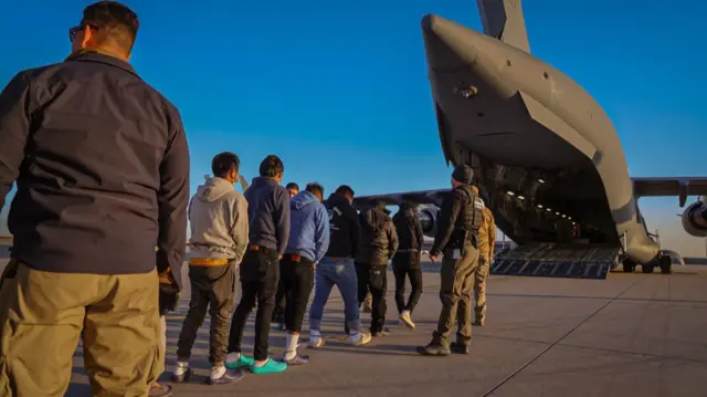 A row of detainees being loaded onto a US military aircraft, aboard a deportation flight