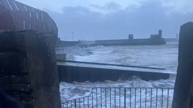 Waves crashing across Whitehaven Harbour seen through a gap in a wall and with a jetty in the foreground and a lighthouse in the far background