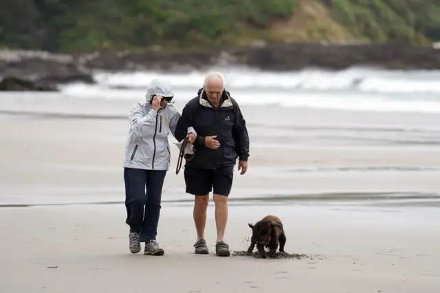 Two people walking their brown dog on a beach. The woman has her hood up and she is holding onto the man's arm.