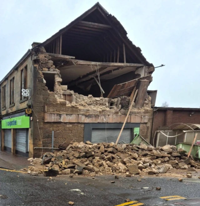 Half a wall is upright, but the top half is in bricks on the road next to it. The inside of the building is visible.