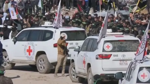 Three Red Cross vehicles drive off after Israeli hostages handed over. A gunman holds a hand up to the second vehicle, large crowds in the background